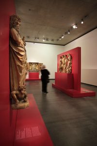 Museumsdesign. Besucher vor Ausstellungswand alten Holzfiguren, Holzfigur im Vordergrund.