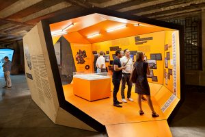 Ausstellungsarchitektur. Begehbarer Ausstellungskörper, Innen, orange. Mehrere Personen stehen vor Abbildungen.