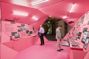 Ausstellungsarchitektur. Begehbarer Ausstellungskörper, Innen, rosa. Zwei Personen stehen an Vitrinen.